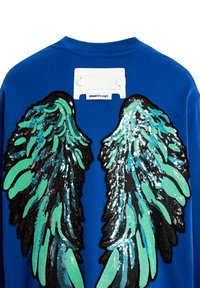 Sudadera azul con grandes diseños de alas en verde y negro, decoradas con lentejuelas, en la parte posterior. Incluye una etiqueta rectangular blanca en el cuello.