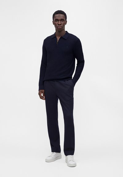 Homme portant un pull bleu marine foncé avec col zippé, pantalon assorti et baskets blanches, debout avec une main dans la poche.