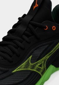 Μαύρο και γκρι αθλητικό παπούτσι με πράσινη σόλα και λογότυπο, μαύρα κορδόνια και πορτοκαλί οφθαλμούς, που δείχνει κοντινή λεπτομέρεια των υφών του πλέγματος και του υφάσματος.