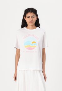 Balta kokvilnas T-kreklu ar apaļu grafiku, kurā ir viļņi, saule un krāsas: zila, rozā un dzeltena. Teksts lasāms "COASTAL WAVES 97 SURF CLUB CA."