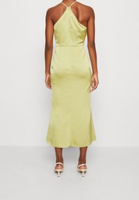 Žena v žlutých saténových midi šatech s tenkými ramínky a zadním zipem, stojící s uvolněnýma rukama podél těla před jednoduchou zdí.