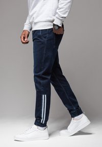 Tummansiniset verkkarit, joissa elastiset resorit lahkeissa, sivuilla kaksi vaaleansinistä pystysuoraa raitaa ja etutaskut. Yhdistetty valkoisten tennareiden kanssa.