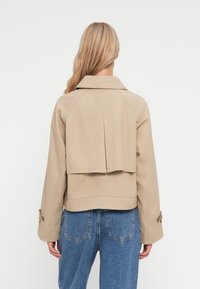 Veste beige avec un design à dos évasé, dotée d'un col et de poignets à boutons. Portée sur un jean en denim bleu. Texture lisse, coupe décontractée.