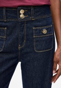 Dark denim jeans with a high-waisted design, featuring two gold buttons at the top, contrast stitching, and front pockets.