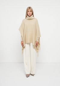 Poncho de tricô bege com gola alta e detalhes de franjas, usado sobre calças claras. Textura suave e silhueta solta e fluida.