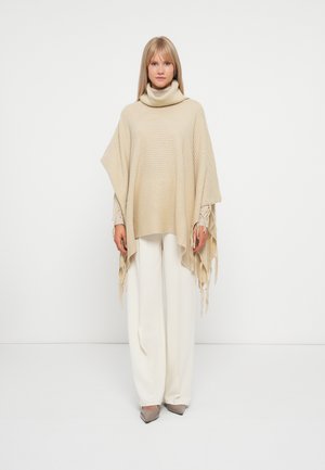 Poncho de tricô bege com gola alta e detalhes de franjas, usado sobre calças claras. Textura suave e silhueta solta e fluida.
