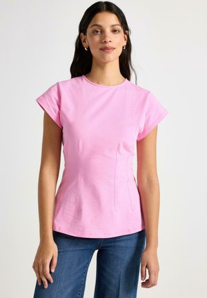 Kvinde med langt mørkt hår iført guldringe i ørerne, en figursyet lyserød kortærmet skjorte og blå jeans mod en ensfarvet baggrund.