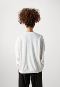 Felpa bianca con vestibilità relaxed, collo a giro e polsini a costine. Tessuto liscio con dettagli di cucitura discreti, abbinata a pantaloni neri a righe.