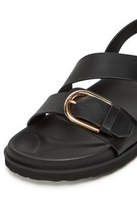 Fekete szandál keresztbe fűzött pántokkal, sima bőrből készült, arany csattal díszítve és texturált talppal a jobb tapadásért.