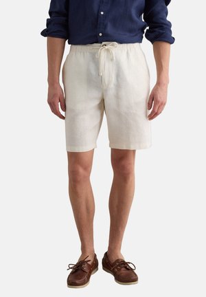 Hombre con pantalones cortos beige con cordón, camisa de manga larga azul marino remangada y zapatos náuticos de cuero marrón, de pie.