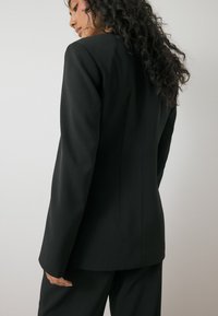 Blazer nero su misura con maniche lunghe, caratterizzato da una texture liscia e opaca e una silhouette strutturata, visto da dietro.