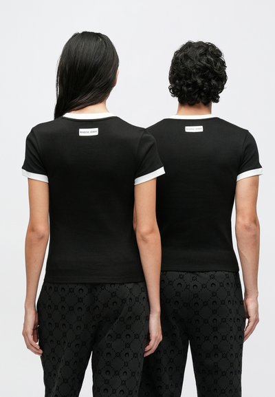 Μαύρα κοντομάνικα t-shirts με λευκή λεπτομέρεια, που φέρουν μια μικρή ετικέτα στο πίσω μέρος. Και οι δύο μοντέλα φορούν μαύρο παντελόνι με μοτίβο που ταιριάζει.