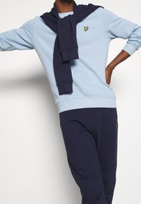 Sudadera azul claro con puños acanalados, combinada con pantalones jogger navy que presentan un pequeño logo amarillo. Suéter azul marino colgado alrededor del cuello.