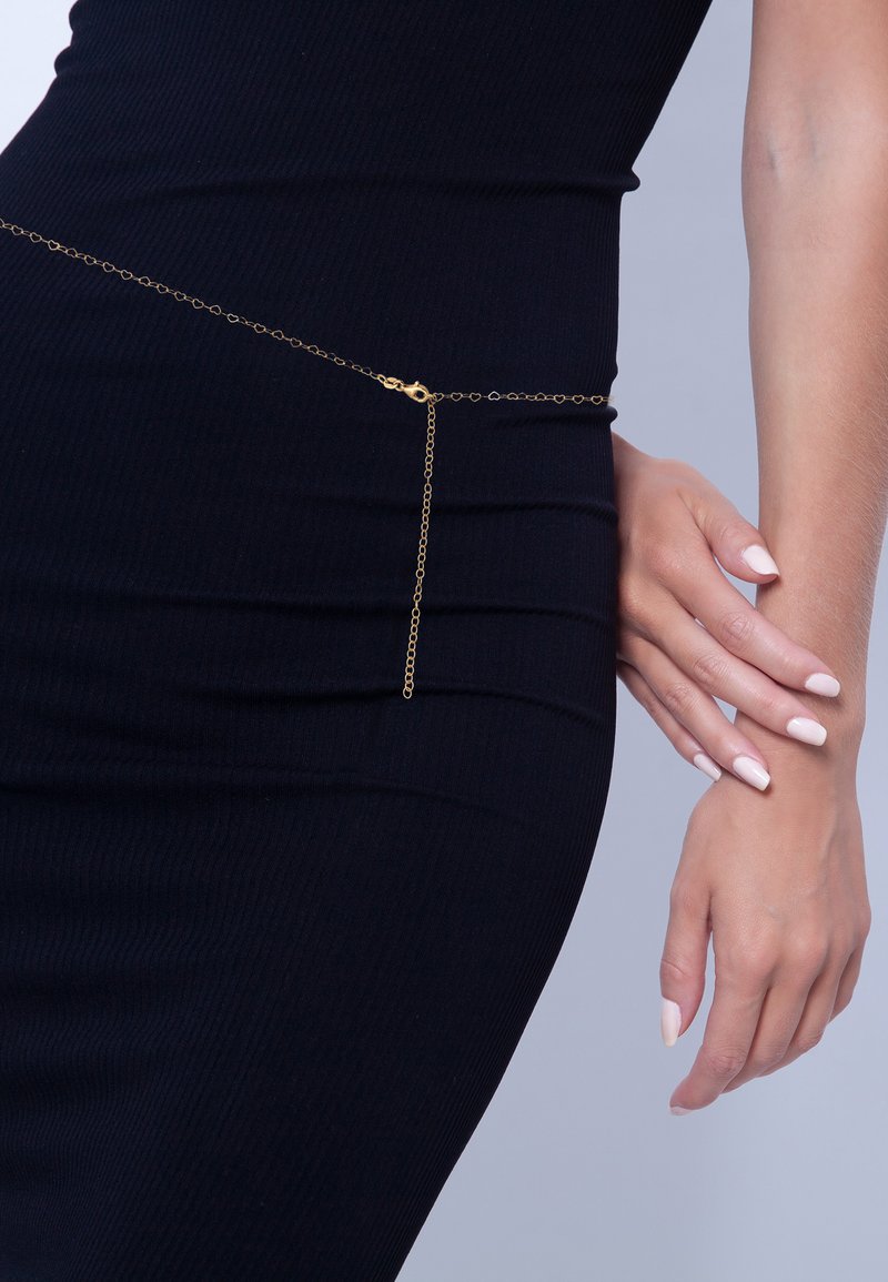 Cintura in catena d'oro con maglie a forma di cuore indossata su un abito nero aderente, con un primo piano della mano e dell'ormo di una donna.