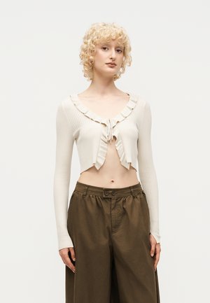Frau mit lockigem blonden Haar, die eine kurze, cremefarbene gerippte Strickjacke mit Rüschen und braune, hoch taillierte Weithosen trägt.