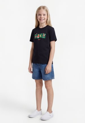 Fată zâmbitoare cu păr lung și blond, purtând un tricou negru Nike cu grafică, pantaloni scurți din denim și pantofi albi slip-on, stând pe un fundal alb.