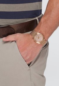 Goldfarbene Uhr mit rundem Ziffernblatt, hellrosafarbenem Zifferblatt und minimalistischen Stundenmarkierungen. Mesh-Metallarmband und ein braunes Lederband im Hintergrund.