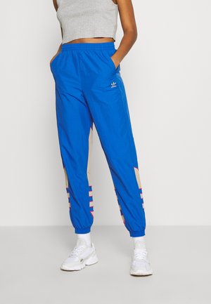 Osoba ubrana w jasnoniebieskie dresy Adidas z beżowymi i różowymi paskami, białe trampki i krótki szary top.