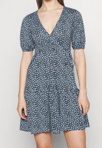 Černé zavinovací šaty s květinovým vzorem v světle modré barvě, s krátkými puffovými rukávy a volánkovým lemem sukně. Vyrobené z lehkého materiálu.