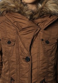 Brun oppustelig jakke med en faux fur hætte, to knap-lukket brystlommer og en lynlås. Den ydre stof er glat med syede detaljer.