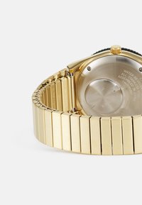 Montre en acier inoxydable doré avec une finition brossée, boîtier rectangulaire, bracelet texturé et un fond transparent exposant les détails mécaniques.