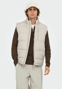 Lys grå polstret vest med høj krave og lynlåslomme, båret over en brun kabelstrikket sweater. Modellen kombinerer det med lyse bukser.