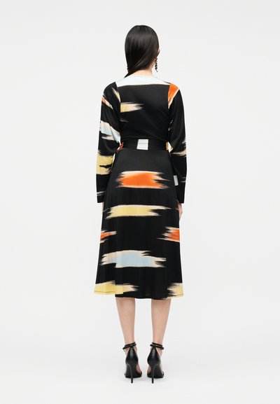 Γυναίκα που φοράει μαύρο φόρεμα μέσης μήκους με μακριά μανίκια και αφηρημένα σχέδια με πινελιές πορτοκαλί, κίτρινου και μπεζ, φωτογραφημένη από πίσω.