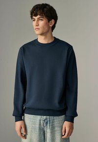 Sudadera azul marino de tejido suave, con cuello redondo y puños acanalados. Combinada con jeans de mezclilla claros y desgastados.
