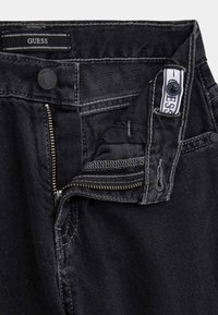 Jeans in denim nero con zip sul fronte, chiusura a bottone e dettagli di cucitura visibili. Etichetta con "GUESS" posizionata all'interno della vita.