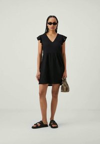 Vestido curto preto com decote em V, mangas rufadas e cintura franzida. Combinado com sandálias pretas de slide e uma pequena bolsa bege.