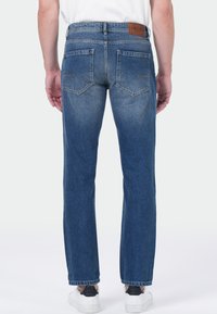 Personne portant un jean en denim bleu coupe droite et des baskets blanches, se tenant de dos face à l'appareil photo sur un fond uni.