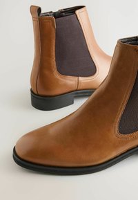 Bottes chevilles en cuir marron avec panneaux latéraux élastiques, bout rond et semelle en caoutchouc noir. Fini lisse avec des détails de coutures sans couture.