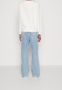 Osoba oblečená v světle modrých širokých cargo džínech a bílém svetru s dlouhým rukávem, stojící zády k fotoaparátu na jednobarevném pozadí.