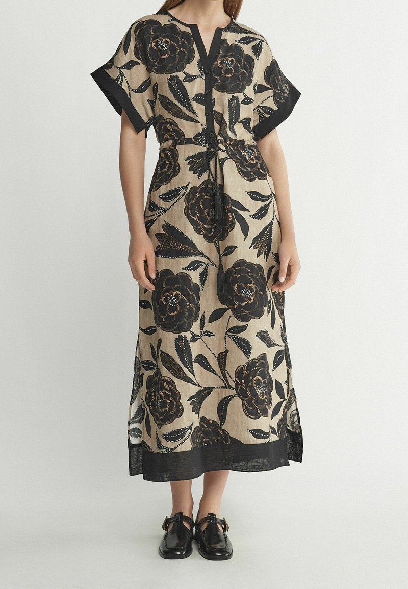 Femme portant une robe midi beige avec un grand imprimé floral noir, manches courtes, devant boutonné et bordure noire aux manches et à l'ourlet, associée à des chaussures noires.