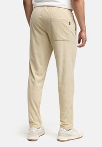Light beige joggers with a ribbed texture, featuring a back pocket. Paired with white sneakers, showing a tailored shape and elastic waistband.