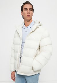 Jeune homme portant un polo bleu clair et une doudoune matelassée blanche avec capuche, se tenant devant un fond blanc uni.