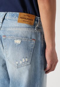 Gai zili denimu džinsi ar nodilušām detaļām, ādas uzlīme ar uzrakstu "JACK & JONES" un klasiskā piecu kabatu dizaina.