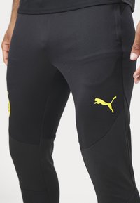 Zwarte sportieve broek van textuurstof met een aansluitend ontwerp, voorzien van een gele Puma-logo op de linker dij en een gladde afwerking.