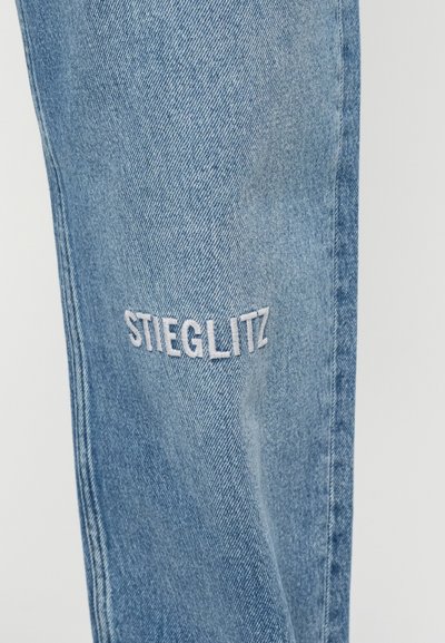 Niebieskie dżinsy z delikatnie zniszczonymi fakturami, z białym haftem "STIEGLITZ" na udzie. Prosty krój nogawki, bez metalowych elementów.