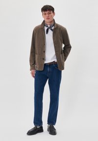 Chaqueta de pana marrón con cuello camisero, cierre de botones, dos bolsillos frontales, llevada sobre una camisa blanca y jeans de mezclilla azul; zapatos negros.