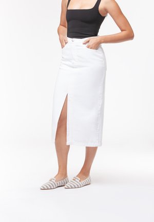 Donna che indossa un top nero senza maniche, gonna midi bianca con spacco frontale e scarpe basse decorate, in piedi con le mani nelle tasche della gonna.