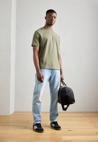 Olive green short-sleeve t-shirt, light blue jeans, black sneakers with white stripes, holding a round black leather bag. Neutral background.