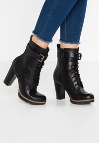 Botas de cuero negras hasta el tobillo con un tacón alto, con cordones en la parte delantera, cremallera lateral y detalle de hebilla en el tobillo. Suela texturizada.