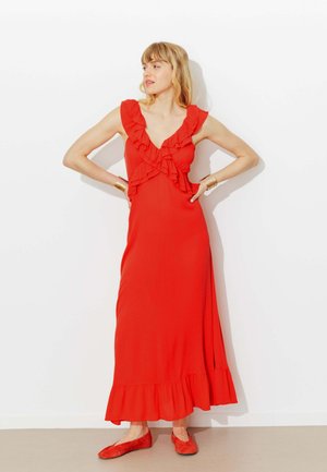 Femme en robe maxi rouge sans manches à volants, posant les mains sur les hanches, portant des ballerines rouges assorties et des bracelets manchettes dorés.