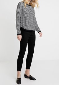 Schwarzer und weißer, strukturierter Pullover mit Rundhalsausschnitt, kombiniert mit enganliegenden schwarzen Hosen und schwarzen Slippern mit goldenen Akzenten.