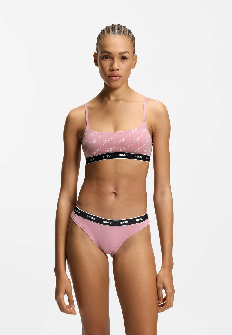 Roze bralette en bikini set met verstelbare bandjes, voorzien van een herhalend wit logo patroon en een zwarte elastische tailleband. Gladde textuur.
