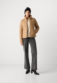 Beige pufferjakke med høj krave, quiltet mønster og lynlås foran. Sammen med grå bredde ben jeans og sorte hæl ankelsko.
