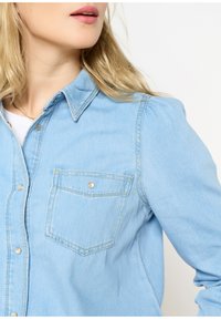 Camicia in denim azzurro chiaro con design a bottoni, tasca sul petto e colletto con dettagli di cucitura sottili.