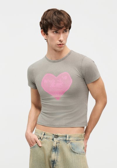 Νέος άνδρας φοράει στενό γκρι κοντό t-shirt με μεγάλο ροζ καρδιά στο κέντρο, σε συνδυασμό με ανοιχτόχρωμο ξεβαμμένο τζιν, χέρια στις τσέπες, κοιτάζοντας πλάγια.