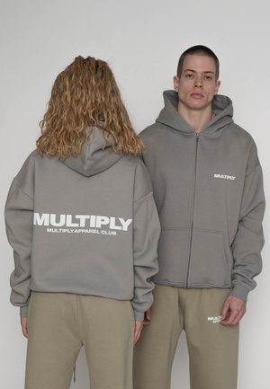 To personer iført grå hættetrøjer og beige bukser, den ene vender fremad, den anden viser teksten "MULTIPLY" på ryggen.